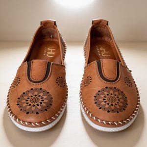 Front view of a pair of handmade tan leather slip-on flats with floral cutout detailing, artisan stitching, and lightweight white soles.
