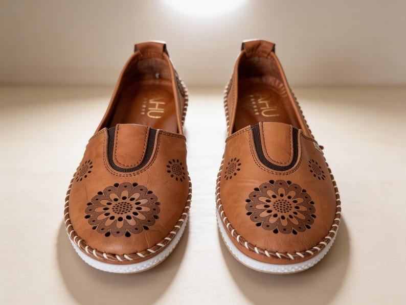 Front view of a pair of handmade tan leather slip-on flats with floral cutout detailing, artisan stitching, and lightweight white soles.