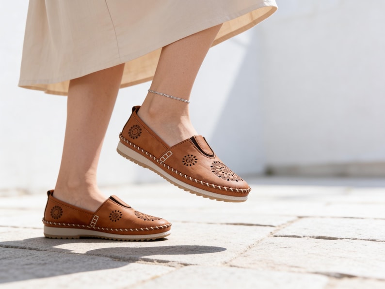 Woman wearing handmade tan leather slip-on flats with floral cutout detailing, walking outdoors in soft sunlight with a cream skirt and silver anklet.