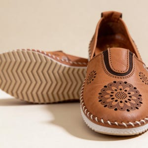 Handmade tan leather slip-on flats with floral cutout detailing, shown from the front with one shoe turned to reveal the textured sole.