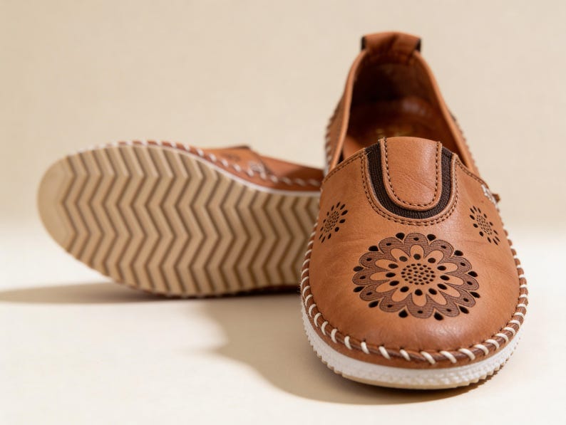 Handmade tan leather slip-on flats with floral cutout detailing, shown from the front with one shoe turned to reveal the textured sole.
