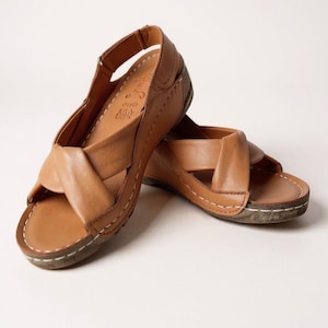 Op de afbeelding: Een paar lichtbruine leren sandalen met een kruisontwerp over de voet. De sandalen hebben een kleine sleehak en witte stiksels rond de randen. De sandalen zijn ongeveer 23 cm lang.