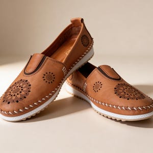 Pair of handmade tan leather slip-on flats with floral cutout detailing and white soles, styled in a crossed position on a soft beige studio background.