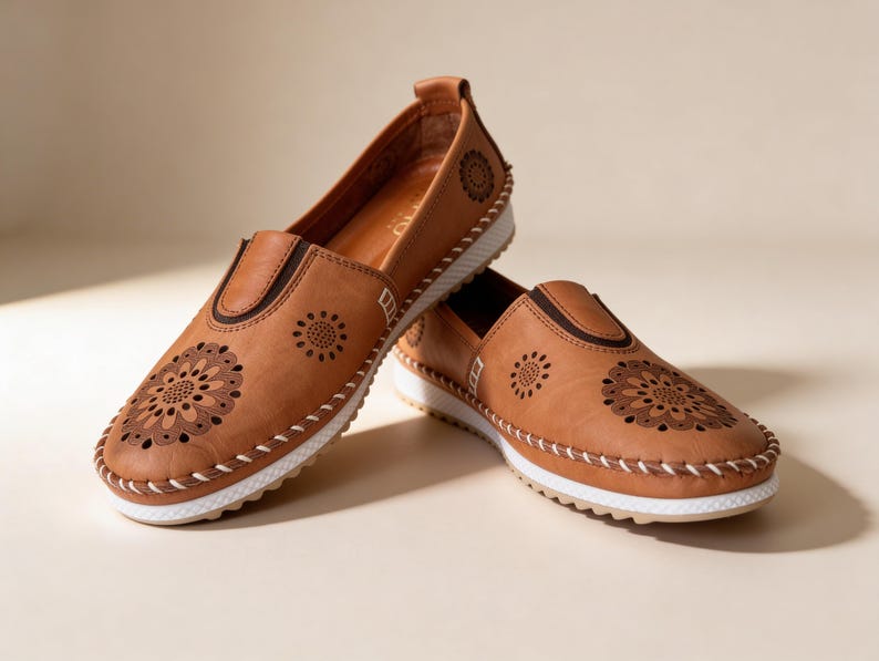 Pair of handmade tan leather slip-on flats with floral cutout detailing and white soles, styled in a crossed position on a soft beige studio background.
