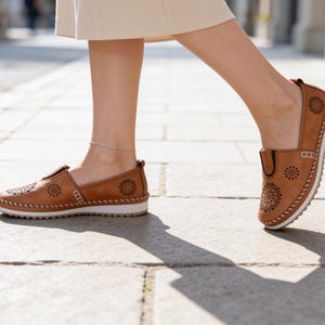 Woman walking outdoors in handmade tan leather slip-on flats with floral cutout detailing, styled with a cream skirt and delicate silver anklet.