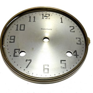 May include: A vintage clock face with a silver dial and a brass bezel. The face has black numerals and the word "Waterbury" printed in the centre. The text "MADE IN U.S.A. BY WATERBURY CLOCK CO." is printed at the bottom.