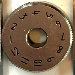 May include: A close-up shot of a metallic dial featuring the numbers 1 to 12 around the edge. The dial has a central aperture and a ridged outer edge. The numbers are in a clear, simple font.