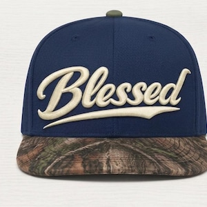 May include: A navy blue baseball cap with a flat brim featuring a brown and green camouflage pattern. The front of the cap displays the word Blessed in large, raised, cream-colored script embroidery. The cap has a small olive green button on top.