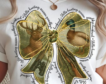 Puede incluir: Camiseta blanca con un diseño en forma de lazo que presenta un ciervo, un pato, unos prismáticos y un rifle. El lazo tiene un centro de camuflaje y ribetes dorados. El texto dice "Hello hunting season goodbye husband."