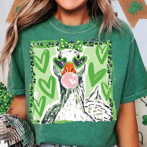 May include: Green t-shirt featuring a whimsical goose design. The goose wears heart-shaped sunglasses and a bow, blowing a bubblegum bubble. The background includes green hearts and a leopard print border.