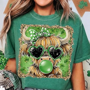 May include: Green t-shirt featuring a graphic of a Highland cow with heart-shaped sunglasses and a green bow. The cow is blowing a bubble and surrounded by shamrocks. The design has a leopard print border, perfect for St. Patrick's Day.