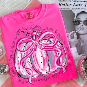 May include: Bright pink Comfort Colors t-shirt featuring a pink pumpkin design with a pink ribbon. The text reads "In October We Wear Pink & Together We Fight." A breast cancer awareness theme.