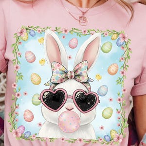 May include: Pink t-shirt featuring a cartoon bunny wearing heart-shaped sunglasses and blowing a bubblegum bubble. The bunny is surrounded by Easter eggs and flowers, with a floral bow and headband. The design has a light blue background.