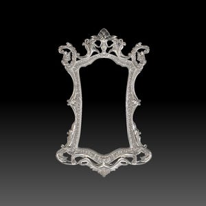 May include: Ornate silver-colored picture frame with a decorative, scrolling design. The frame has a rectangular opening and is set against a black background. The frame's design includes floral and scrollwork details.