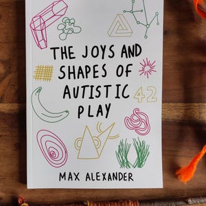 May include: A book titled "THE JOYS AND SHAPES OF AUTISTIC PLAY" by Max Alexander. The cover is white with colorful line drawings of shapes and a banana. The number 42 is also on the cover. The book is on a wooden surface.