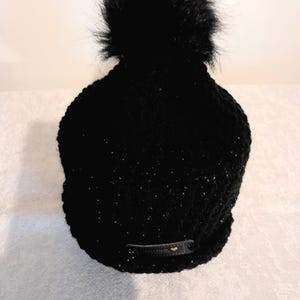 May include: A black knit beanie with a large, fluffy black pom-pom on top. The hat has a subtle sparkle and a small, dark-colored tag near the brim. This winter hat is a stylish accessory.