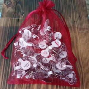 Pode incluir: Uma bolsa de malha vermelha cheia de vários anéis prateados. Cada anel tem uma etiqueta branca com o texto "Stainless steel" e um número de tamanho. A bolsa é amarrada com uma fita vermelha e está sobre madeira.