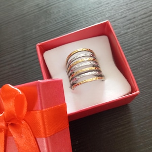 Pode incluir: Um anel de várias faixas com detalhes dourados, prateados e vermelhos, apresentado numa caixa de presente vermelha com uma fita a condizer. O anel apresenta um design texturizado.