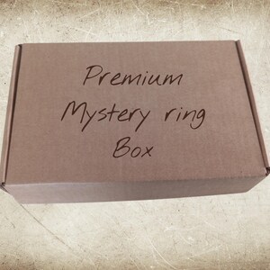 Pode incluir: Uma caixa de cartão castanha com as palavras "Premium Mystery ring Box" escritas à mão. A caixa retangular tem um design simples, sugerindo um presente ou uma caixa de assinatura.