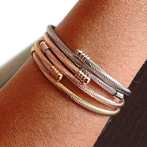 Może przedstawiać: Kolekcja czterech bransoletek w stylu kablowym w odcieniach złota, różowego złota i srebra. Bransoletki są ułożone jedna na drugiej, prezentując ich teksturowany design i dekoracyjne zakończenia. Biżuteria jest prezentowana na ramieniu.