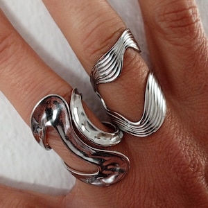 Può includere: Due anelli d'argento sono mostrati su un dito. Gli anelli hanno un design unico e fluido. Un anello presenta una serie di linee parallele, mentre l'altro ha una forma più organica e curva. Gli anelli sono molto lucidi e riflettono la luce.