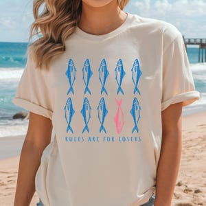 May include: Cream-colored t-shirt featuring a pattern of blue fish, with one pink fish, and the text "RULES ARE FOR LOSERS" in blue. The shirt has short sleeves and a relaxed fit.