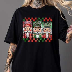 May include: Black t-shirt featuring a colorful graphic of three nutcracker figures wearing crowns. The nutcrackers have white mustaches and are set against a red and green checkered background. The design is festive and holiday-themed.