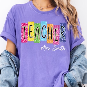 May include: A purple t-shirt with the word "TEACHER" in black letters with a colorful background. Below the word "TEACHER" is the name "Mrs. Smith" in white script. The shirt has rolled-up sleeves.
