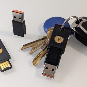 May include: A black USB security key with an orange accent is attached to a keyring with keys and a car key fob. Another key is visible in the background. The key has a gold "y" logo.