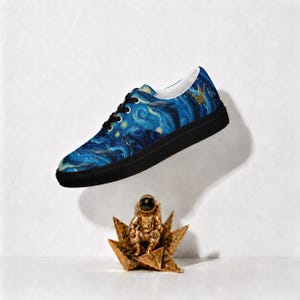 May include: A low-top sneaker featuring a blue and yellow swirling pattern reminiscent of a starry night sky, black laces, and a solid black rubber sole. Below the shoe sits a small, metallic gold astronaut figurine on a star-shaped base.