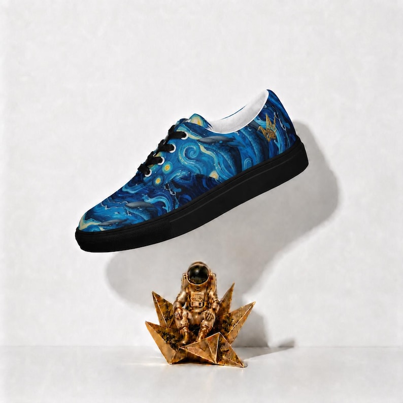 May include: A low-top sneaker featuring a blue and yellow swirling pattern reminiscent of a starry night sky, black laces, and a solid black rubber sole. Below the shoe sits a small, metallic gold astronaut figurine on a star-shaped base.
