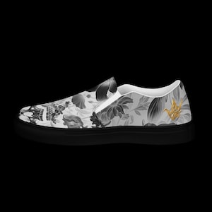 May include: A slip-on sneaker featuring a grayscale floral and skull pattern across the upper canvas. The shoe has a solid black rubber sole and a small, metallic gold origami crane emblem positioned near the heel on the outer side.