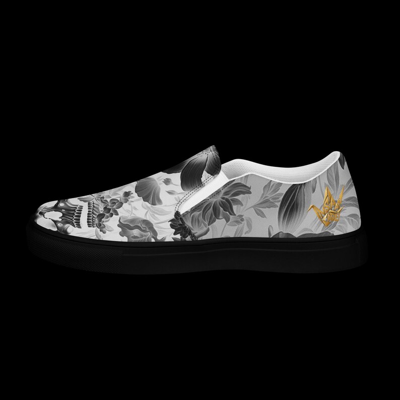 May include: A slip-on sneaker featuring a grayscale floral and skull pattern across the upper canvas. The shoe has a solid black rubber sole and a small, metallic gold origami crane emblem positioned near the heel on the outer side.