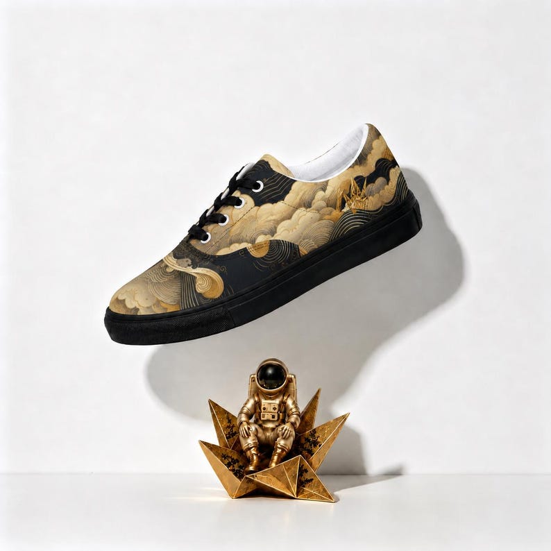 May include: A low-top sneaker featuring a black sole and black laces, with an upper patterned in gold and black swirling cloud designs. Below the sneaker sits a small gold-colored astronaut figurine perched on a geometric star-shaped base.