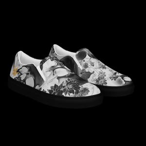 May include: A pair of slip-on sneakers featuring a black and white floral and abstract pattern across the upper fabric. The shoes have solid black rubber soles and a small gold-colored emblem on the outer side of the heel area.