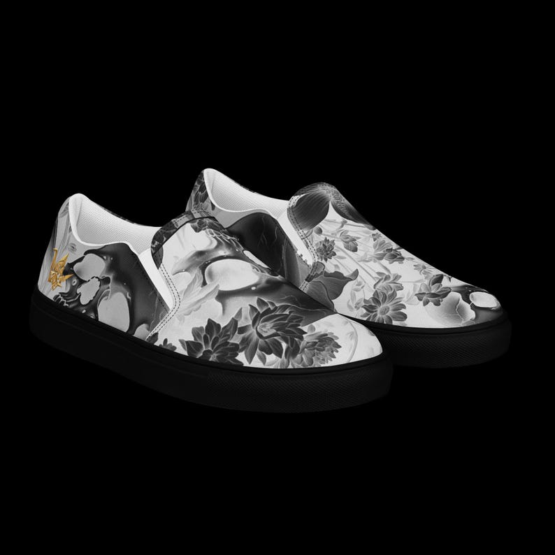 May include: A pair of slip-on sneakers featuring a black and white floral and abstract pattern across the upper fabric. The shoes have solid black rubber soles and a small gold-colored emblem on the outer side of the heel area.