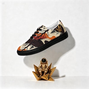 May include: A low-top sneaker with a black sole and black laces, featuring a cream-colored upper with a graphic print of dark brown trees, orange foliage, and black ferns. Below the shoe sits a small gold astronaut figurine on a gold star base.