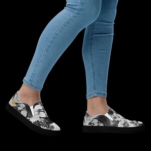 May include: A pair of slip-on sneakers featuring a black and white graphic print of skulls and floral patterns, paired with light blue denim jeans. The shoes have a solid black sole and a low-top design with white elastic side panels for fit.
