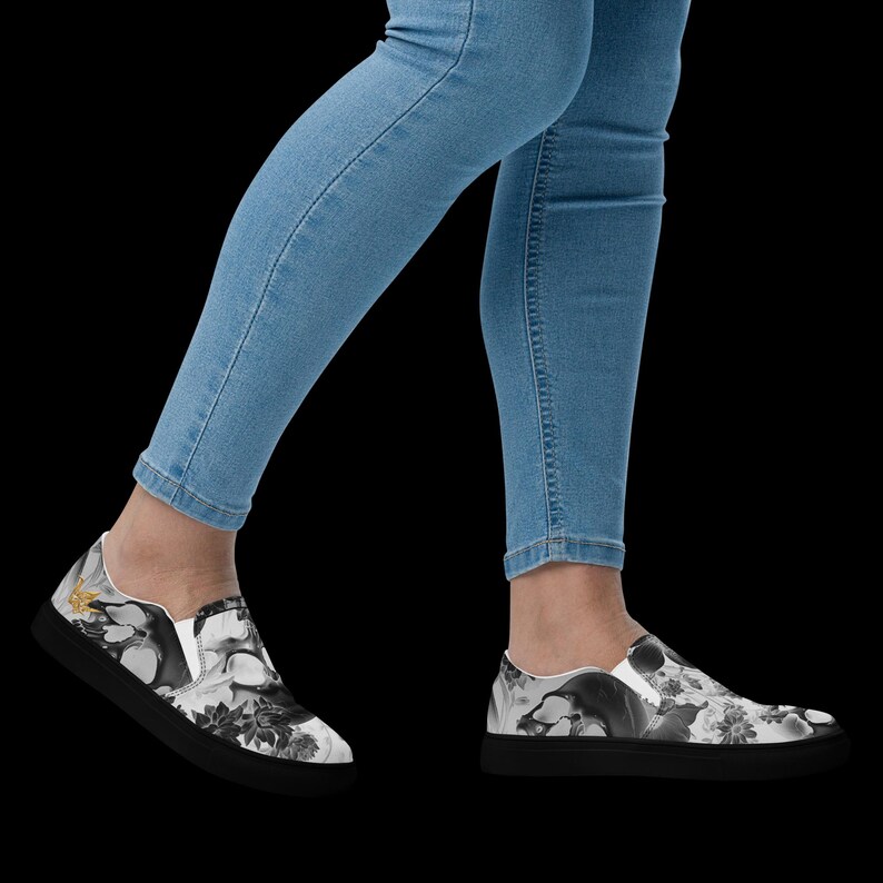 May include: A pair of slip-on sneakers featuring a black and white graphic print of skulls and floral patterns, paired with light blue denim jeans. The shoes have a solid black sole and a low-top design with white elastic side panels for fit.