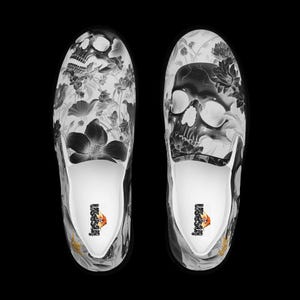 May include: A pair of slip-on sneakers featuring a black and white graphic print of skulls and various floral patterns. The interior of the shoes is white with a small logo printed on the heel area. The design covers the entire upper surface.