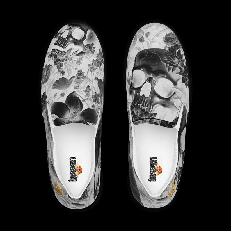 May include: A pair of slip-on sneakers featuring a black and white graphic print of skulls and various floral patterns. The interior of the shoes is white with a small logo printed on the heel area. The design covers the entire upper surface.