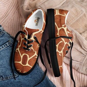 May include: A pair of low-top sneakers featuring a brown and cream giraffe-inspired pattern. The shoes have black laces, black soles, and a white interior lining with a small logo printed on the insole. The exterior fabric displays a repeating giraffe print.