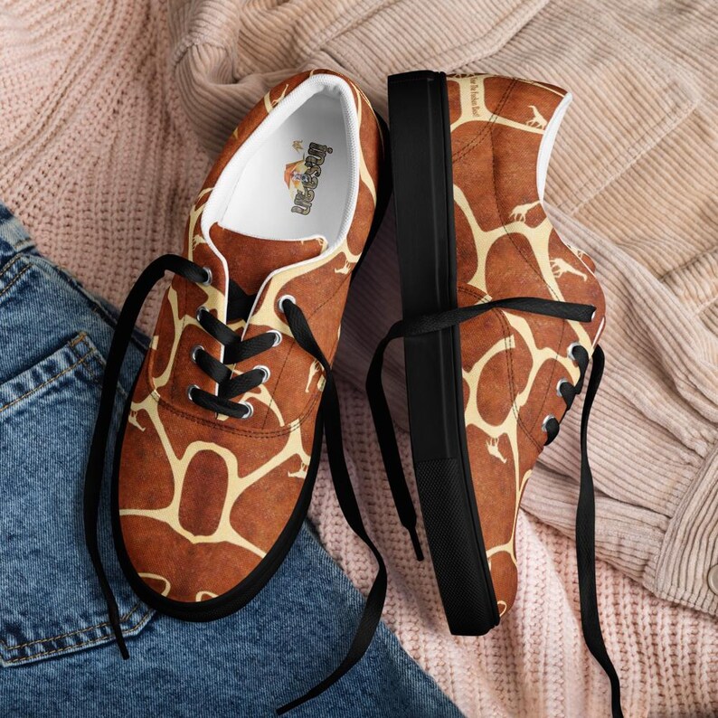 May include: A pair of low-top sneakers featuring a brown and cream giraffe-inspired pattern. The shoes have black laces, black soles, and a white interior lining with a small logo printed on the insole. The exterior fabric displays a repeating giraffe print.