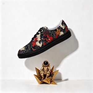 Könnte beinhalten: Ein Low-Top-Sneaker mit schwarzer Basis und einem sich wiederholenden Muster aus stilisierten roten, weißen und schwarzen Wolken. Der Schuh hat schwarze Schnürsenkel, eine schwarze Sohle und ein weißes Innenfutter. Darunter steht eine goldene Astronautenfigur.