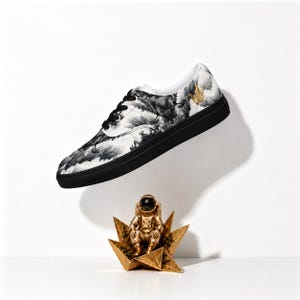 May include: A low-top sneaker with a black sole and a white upper featuring a black and white ink-wash style landscape print and a small gold crane detail. Below the shoe sits a gold-colored astronaut figurine perched on a gold star-shaped base.
