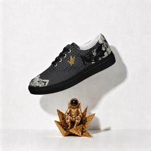 May include: A low-top sneaker with a black sole and a dark grey upper featuring a repeating wave pattern, a gold crane emblem, and white graphic illustrations of figures. Below the shoe sits a small gold astronaut figurine on a star-shaped base.