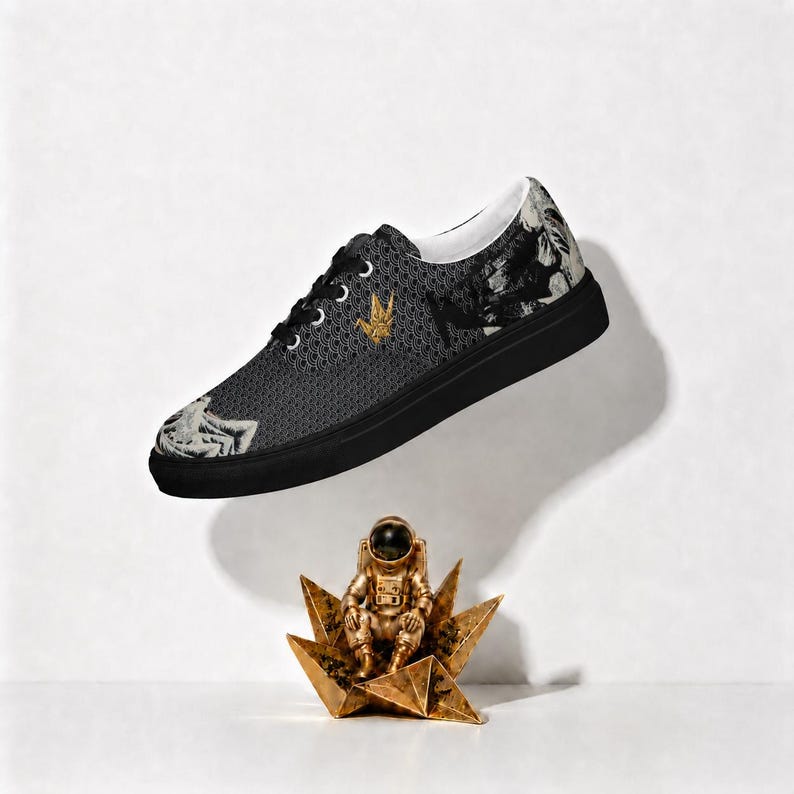 May include: A low-top sneaker with a black sole and a dark grey upper featuring a repeating wave pattern, a gold crane emblem, and white graphic illustrations of figures. Below the shoe sits a small gold astronaut figurine on a star-shaped base.