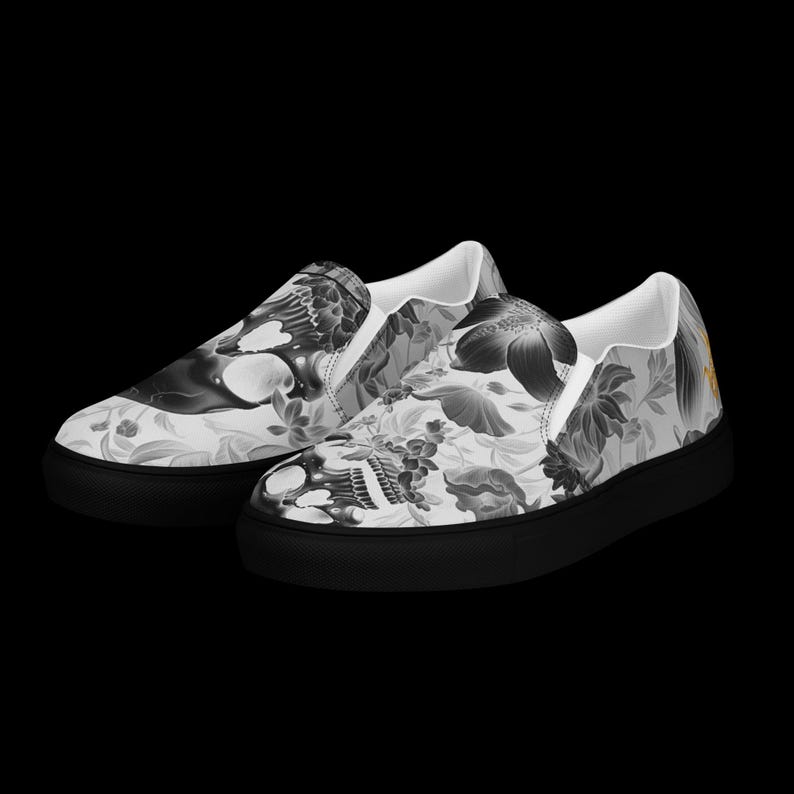 May include: A pair of slip-on sneakers featuring a monochromatic grayscale pattern of skulls and various floral elements across the upper fabric. The shoes have a solid black rubber sole and a white interior lining, providing a stark contrast.