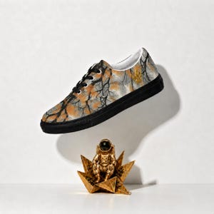 May include: A low-top sneaker featuring a black and orange tree branch pattern on the upper, black laces, and a solid black sole. A small gold emblem is visible on the side. Below the shoe sits a gold-colored astronaut figurine on a star base.