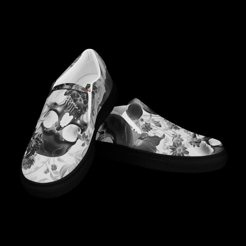 May include: A pair of slip-on sneakers featuring a black and white graphic print of a skull surrounded by various floral patterns. The shoes have a solid black rubber sole and a white interior lining, showcasing a detailed monochromatic design.